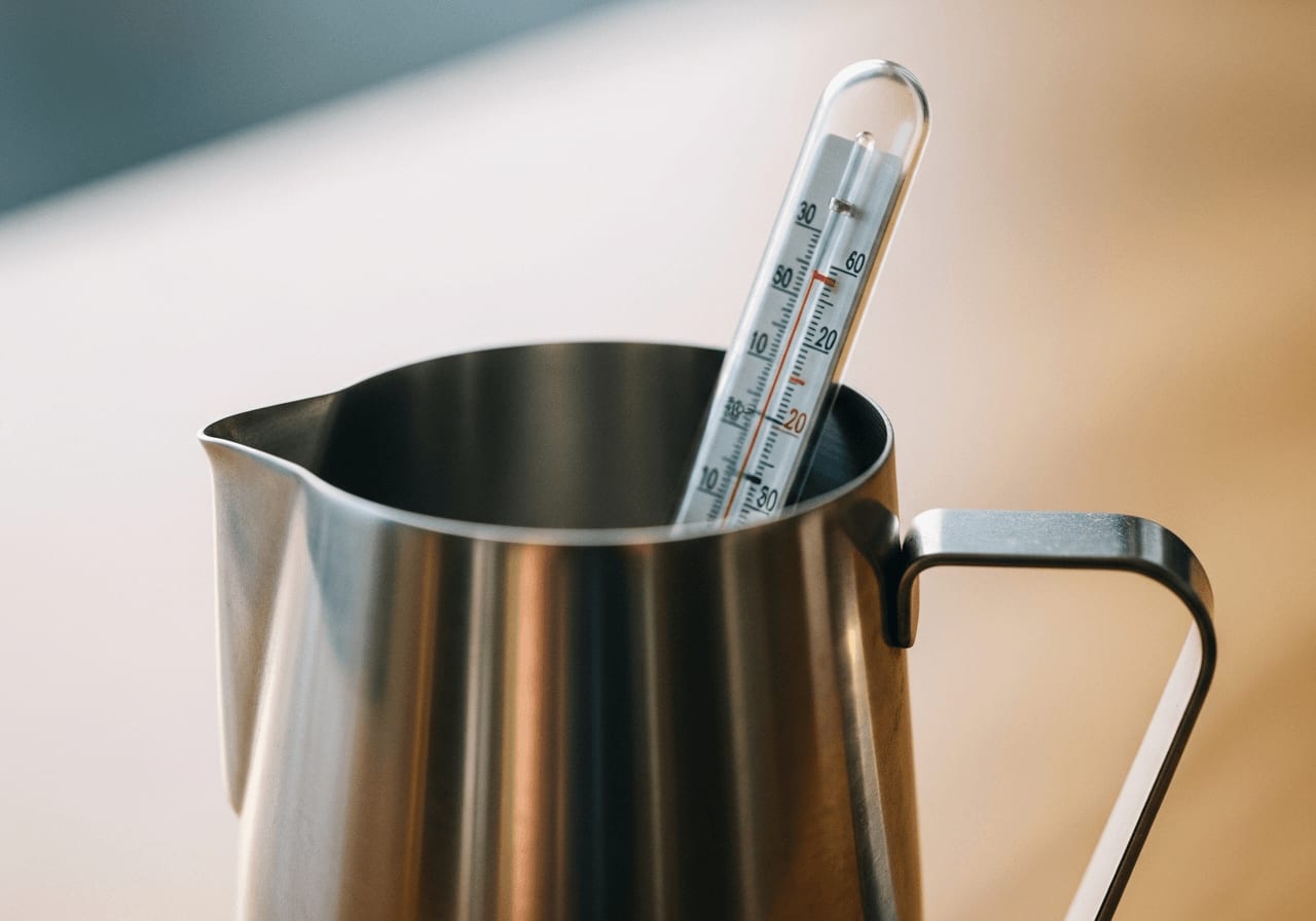 Thermometer in einem Edelstahl-Milchkrug, das die Temperatur der Milch misst, Nahaufnahme